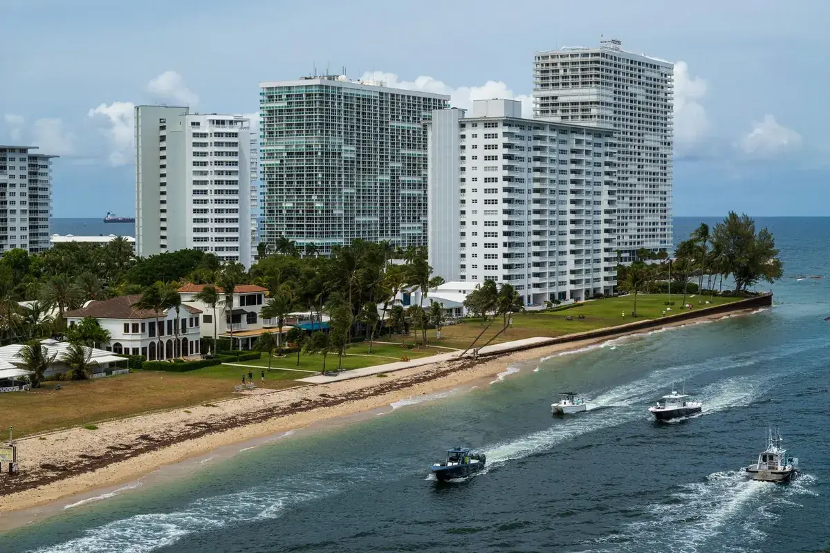 White high-rise condominium towers with balconies beside an inlet and beach, with boats on the water