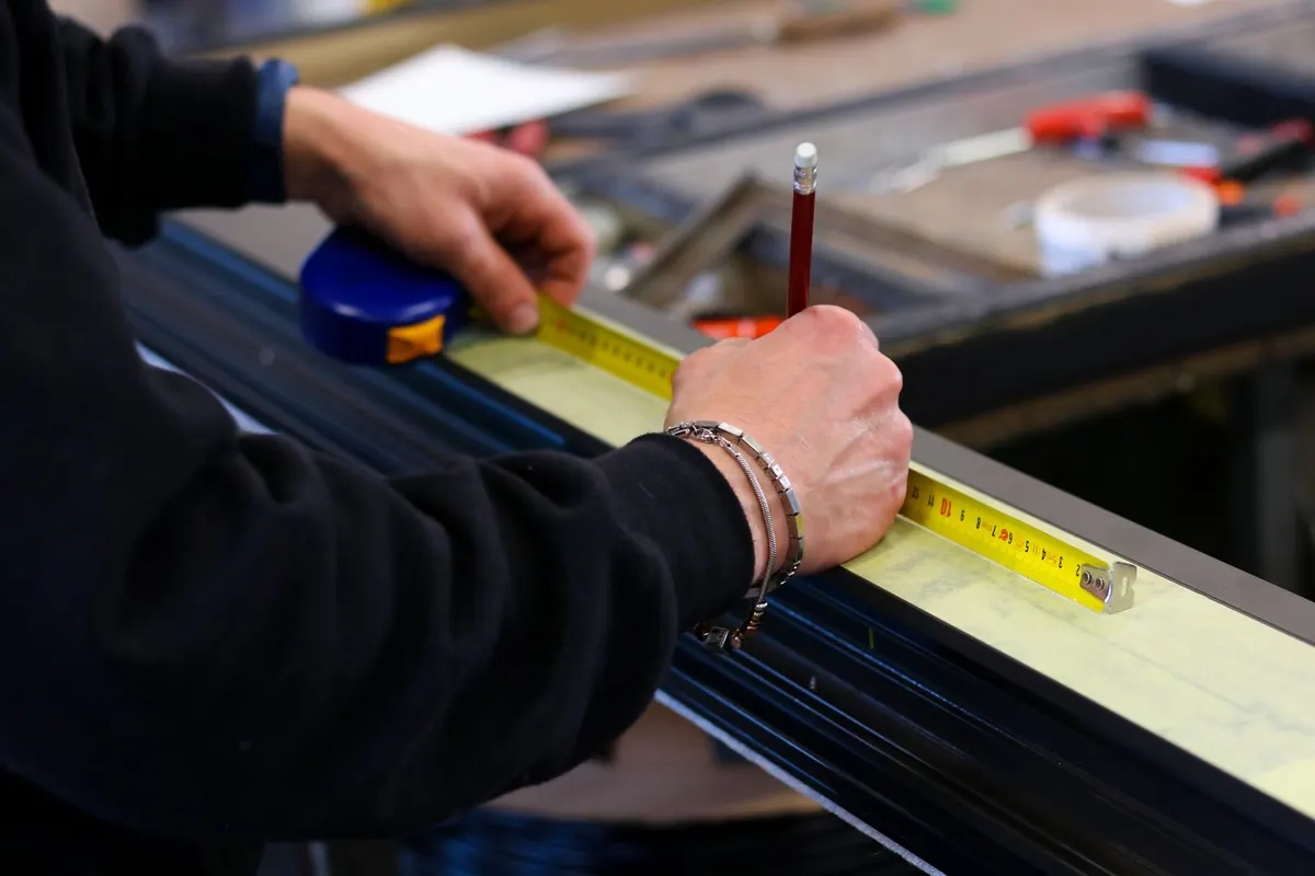 Hands holding a yellow tape measure along an aluminum window profile and marking it with a pencil