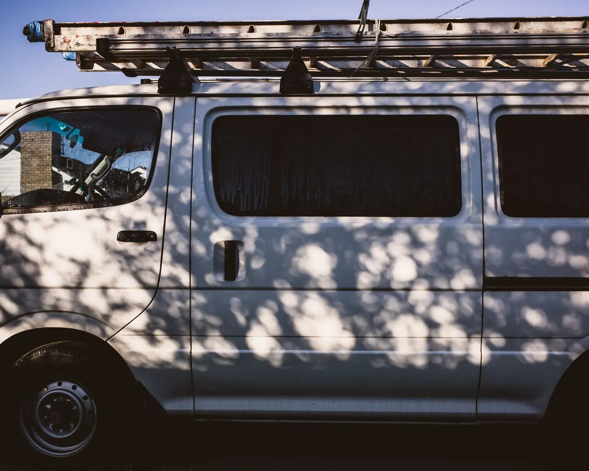 Plain white work van with ladders on the roof rack, parked under trees