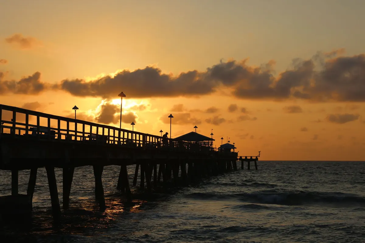 Wooden fishing pier with lamp posts silhouetted against an orange sunrise over the ocean