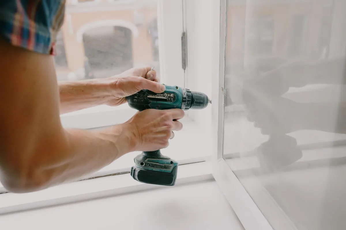 Hands driving a screw into a white window frame with a cordless drill
